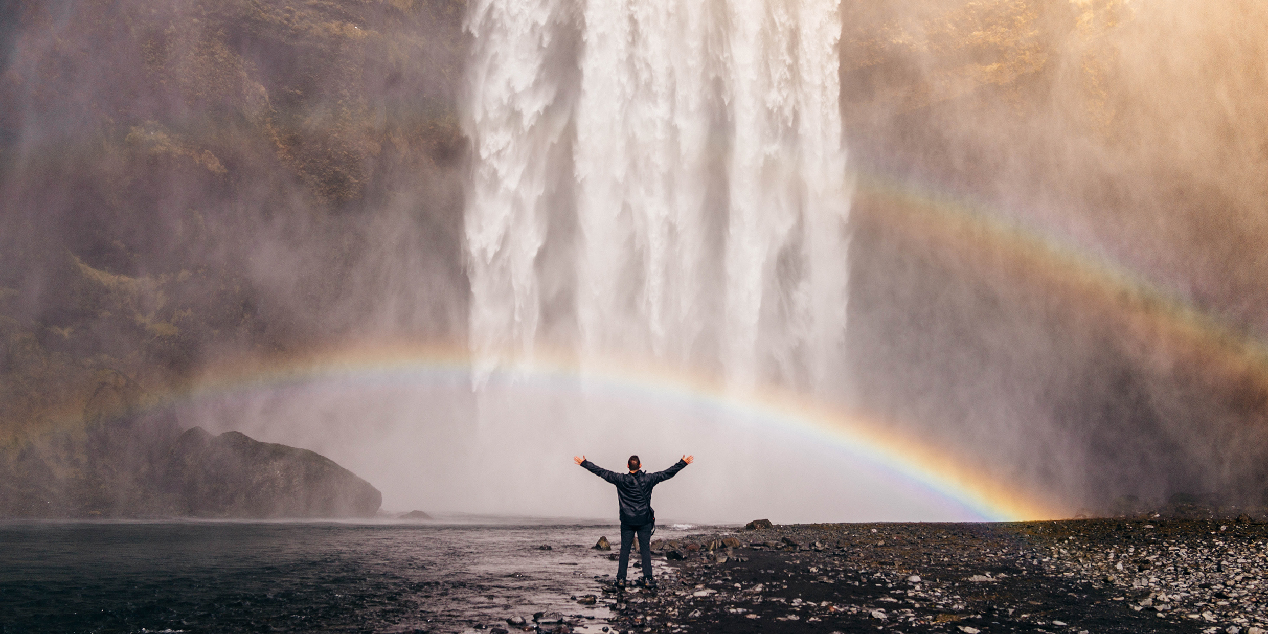Image of man with outstretched arms in front of rainbow created by spray of waterfall