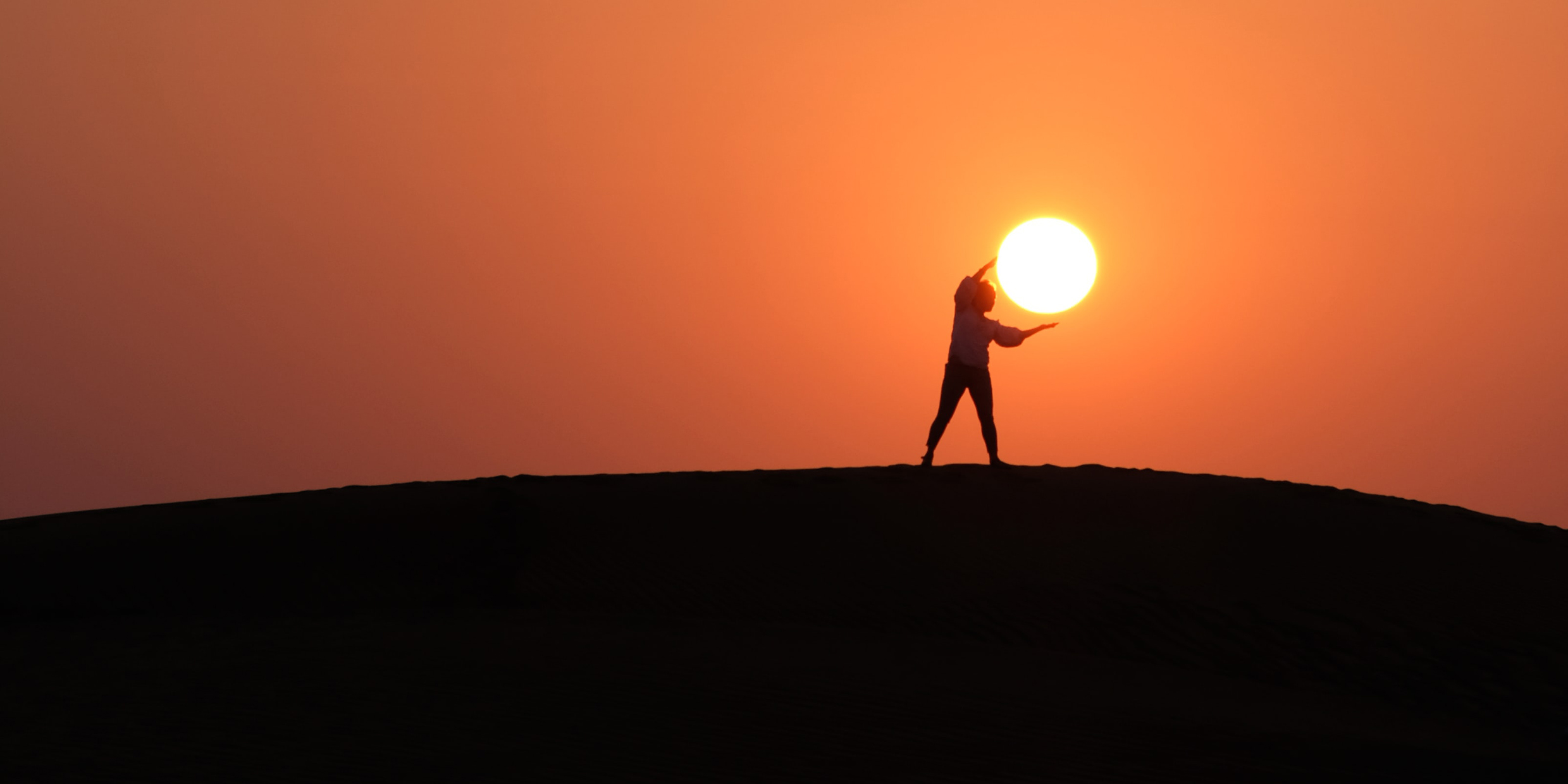 Silhouette of a person standing on the horizon positioned in such a way that they appear to be holding the sun in their hands