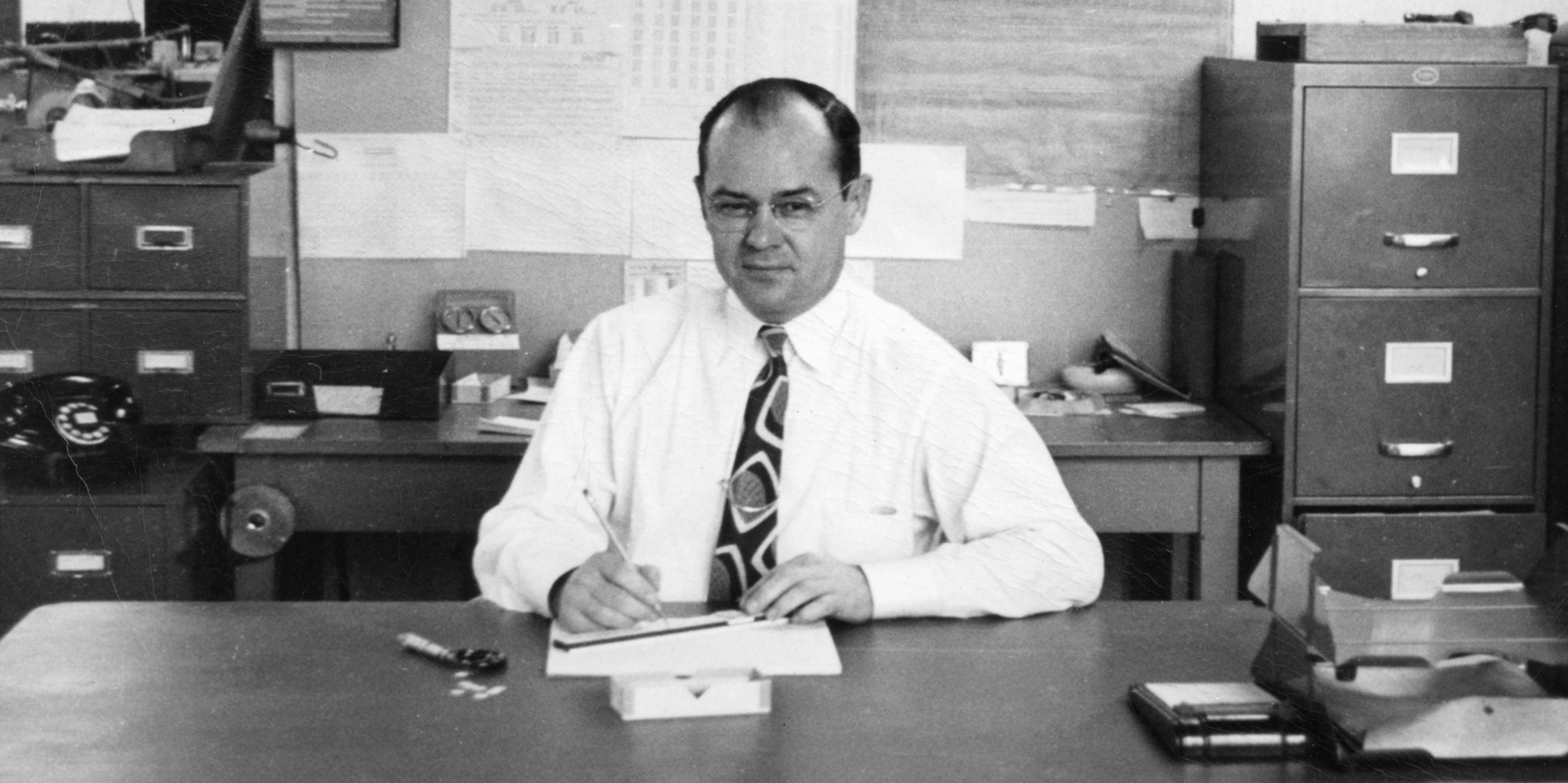 Black and white photo of a man in shirt and tie at a desk with pencil and slide rule