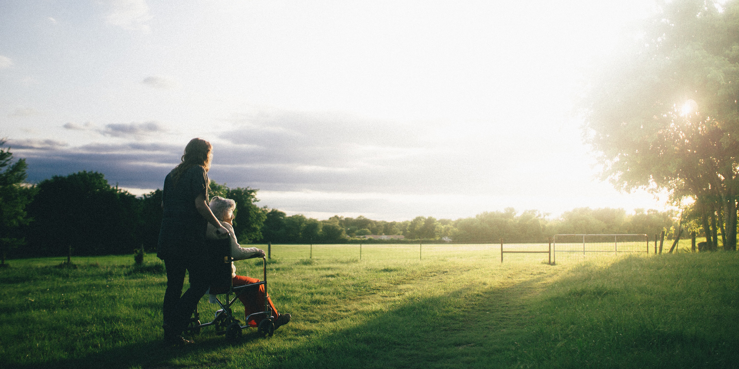 Image of a woman pushing an elderly woman in a wheelchair toward a sunset